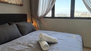 A neatly made bed with a rolled towel, a headboard, and a city view through a window with curtains. - Cozy 2BR Sky Pool & Panoramic Views 1-6 Guests (Gelugor)