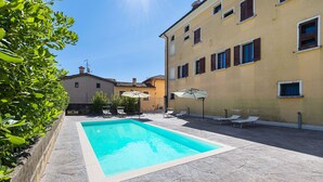 A swimming pool with lounge chairs and umbrellas, surrounded by a stone patio and a building with multiple windows. - Spacious 2 bedroom apartment with pool (Soiano del Lago)