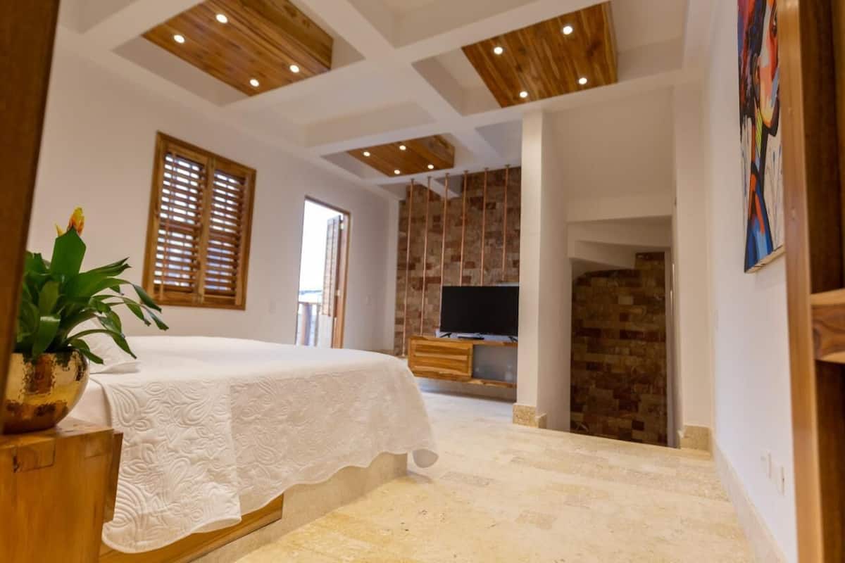 A bedroom with a bed, wooden ceiling beams, a TV, and a stone wall.