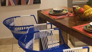 Un coin repas avec une table en bois, des chaises bleues, un tableau coloré, et un espace cuisine avec des étagères et des appareils électroménagers.