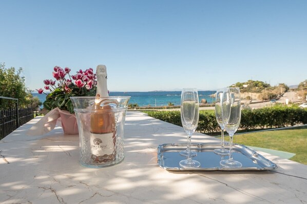 Une table avec une bouteille de vin rosé Amica, deux verres et un arrangement floral, le tout placé devant une vue panoramique sur la côte.