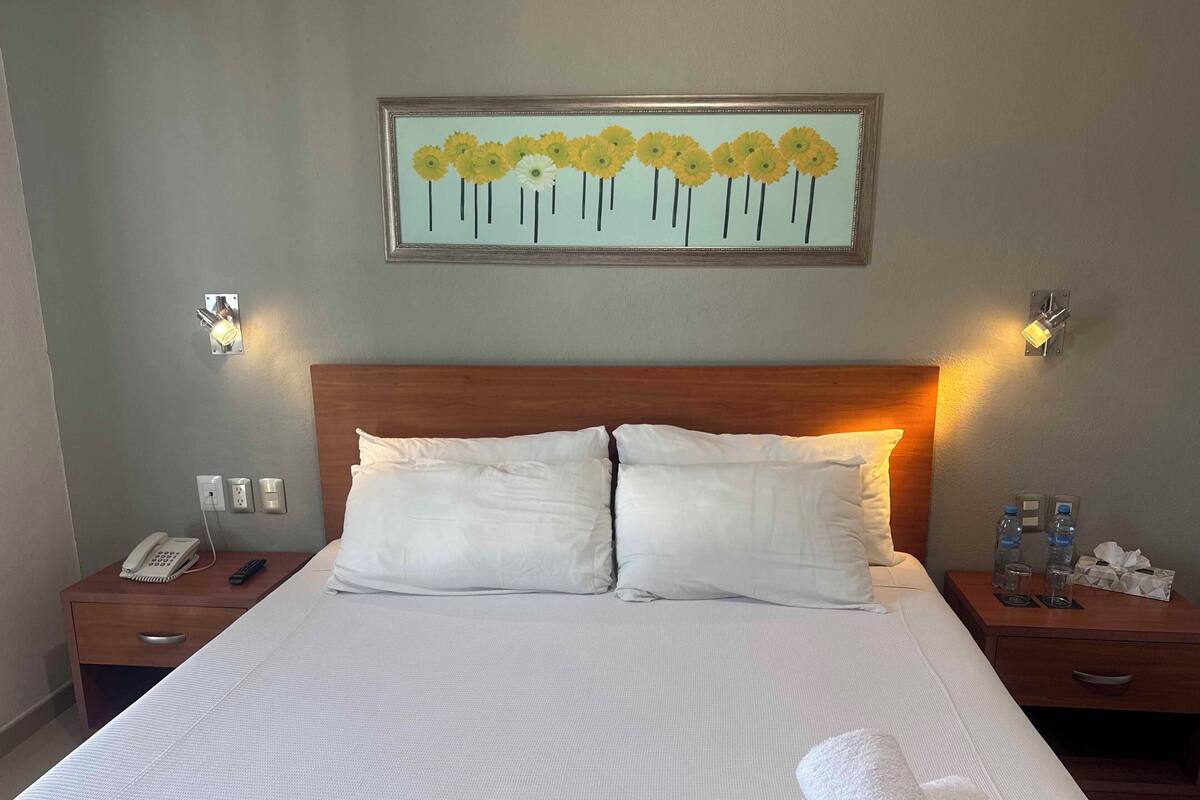 Una cama bien hecha con ropa blanca, un cabecero de madera, mesitas de noche con lámparas y una obra de arte floral enmarcada en la pared.