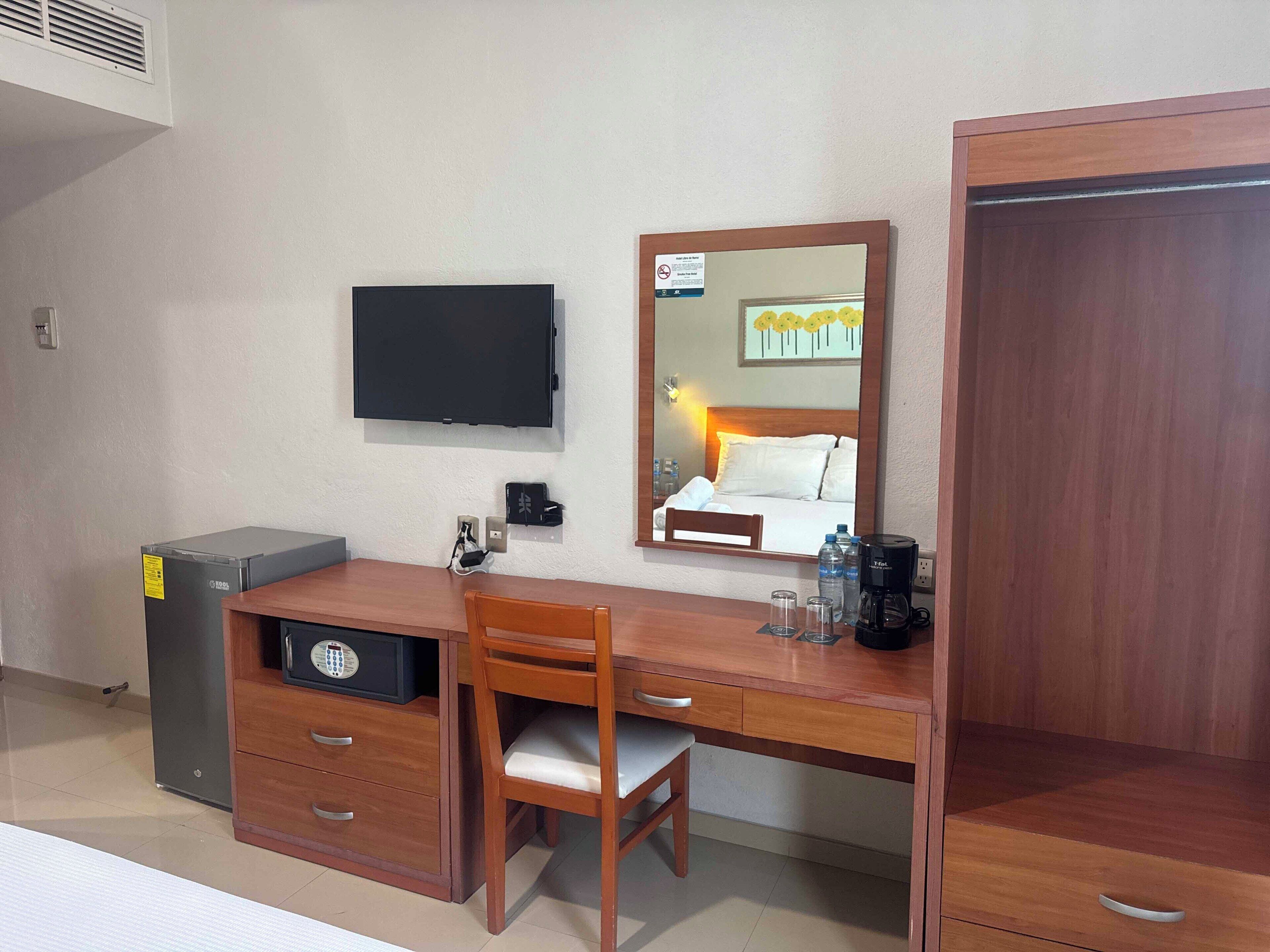 Ein Hotelzimmer mit einem Holztisch, einem Sessel, einem Fernseher, einem Spiegel, einem Kleiderschrank und einer kleinen Mülltonne.
