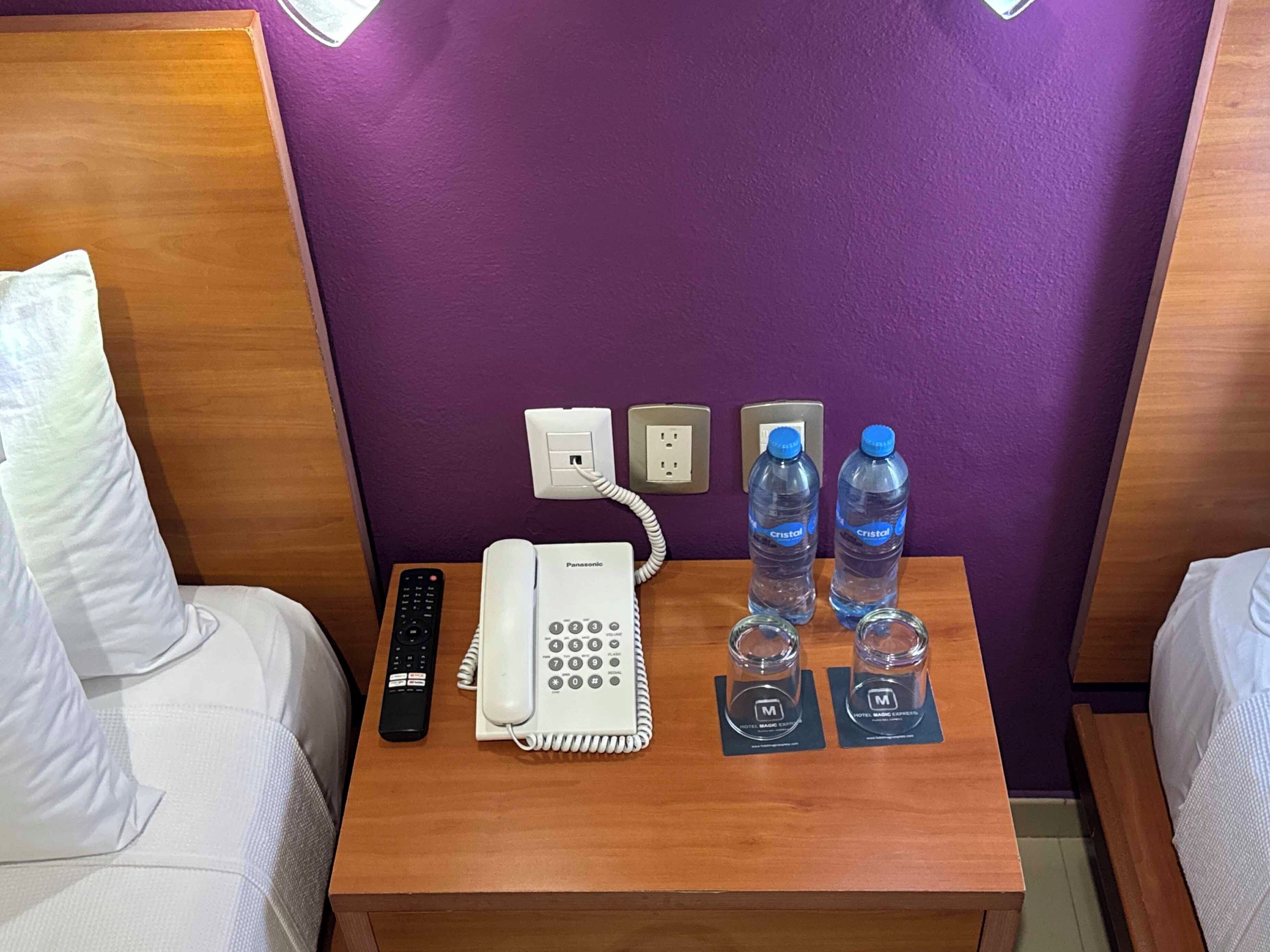 A hotel room with a wooden nightstand, a white phone, a remote control, two Cristal water bottles, and a purple wall.