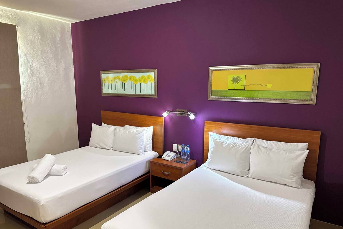 Una habitación de hotel con dos camas, ropa de cama blanca, un cabecero de madera, una mesita de noche con teléfono y botellas de agua, y dos cuadros en la pared.