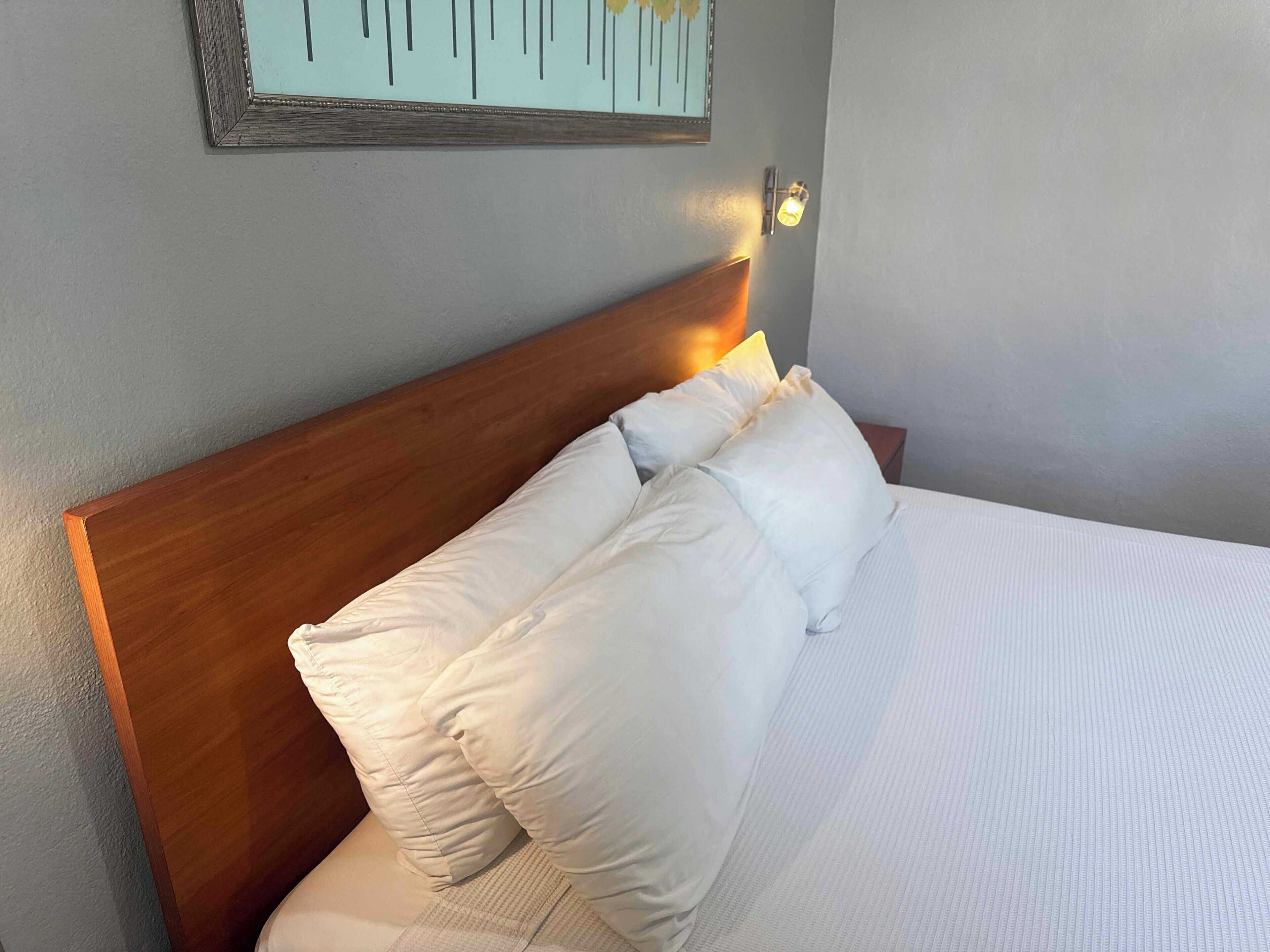 A neatly made bed with white linens and pillows, a wooden headboard, and a wall-mounted light fixture.