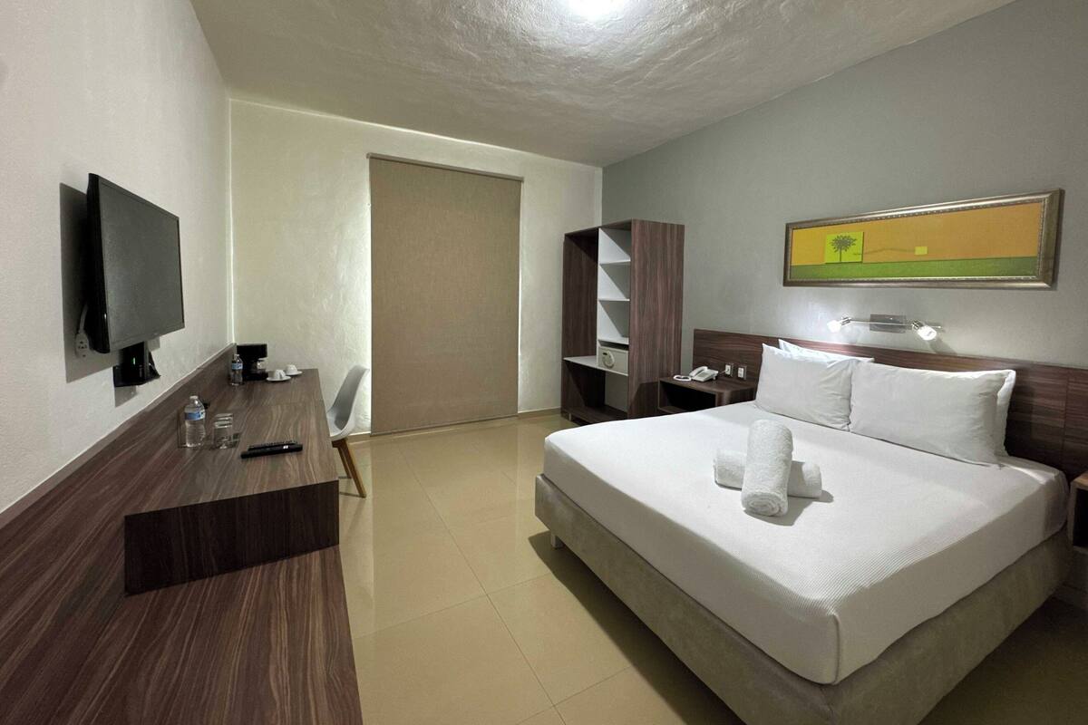 Habitación de hotel con cama, escritorio con televisor de pantalla plana, estantería y un cuadro en la pared.