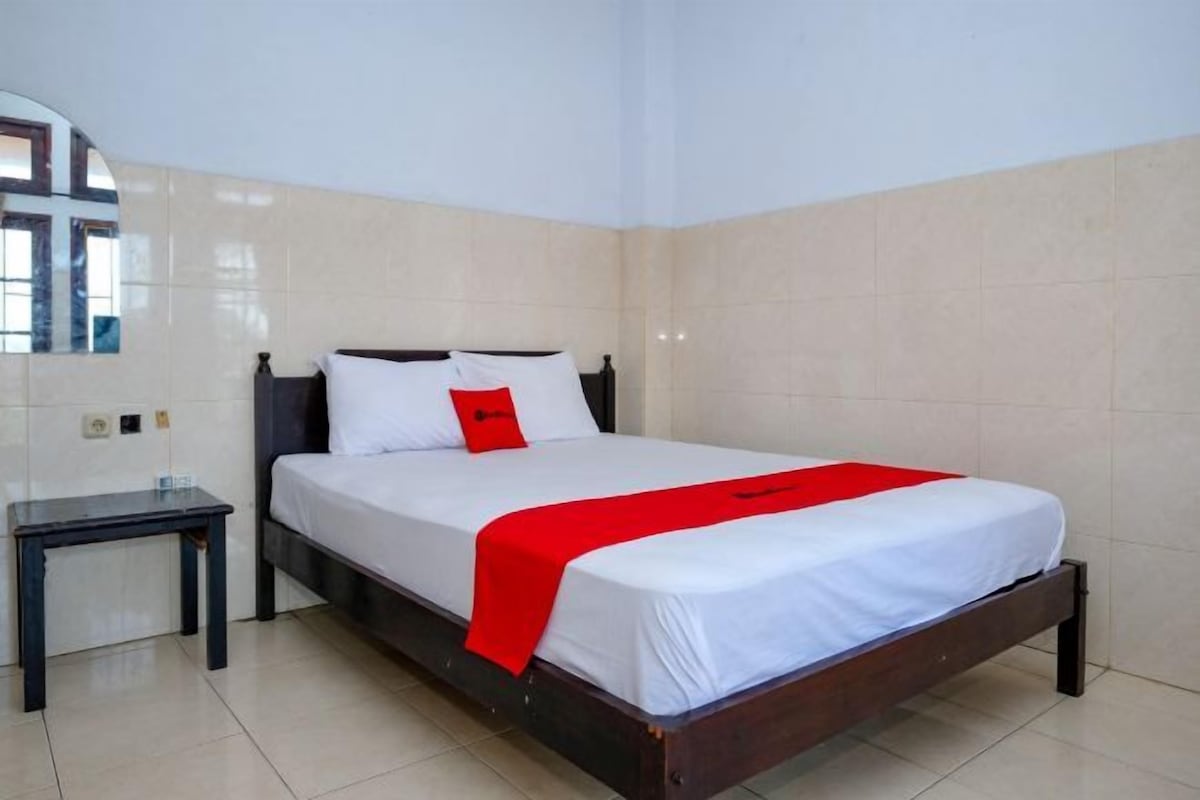 Una cama individual con estructura de madera, ropa de cama blanca y una almohada roja de adorno.