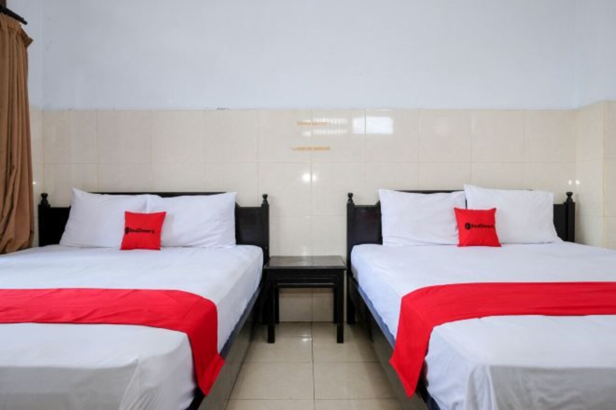 Habitación de hotel con dos camas, cada una con ropa de cama blanca y detalles en rojo, mesitas de noche pequeñas y pared de azulejos.