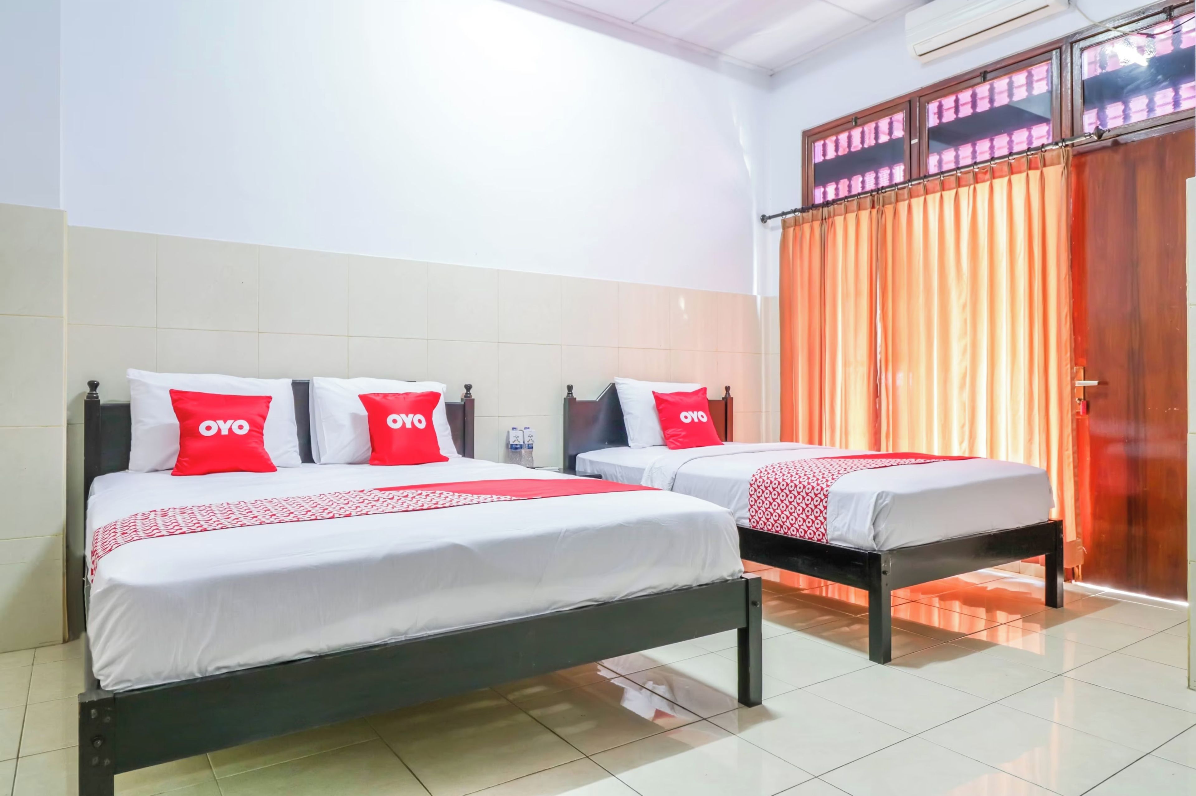 A hotel room with two beds, each with white bedding and red pillows, and a large window with orange curtains.