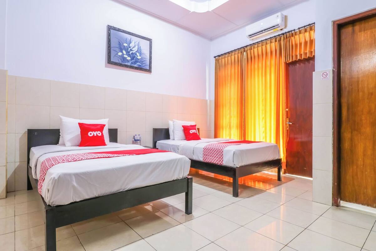 A hotel room with two beds, each with white bedding and red pillows, a framed floral artwork on the wall, and an air conditioning unit above the bed.