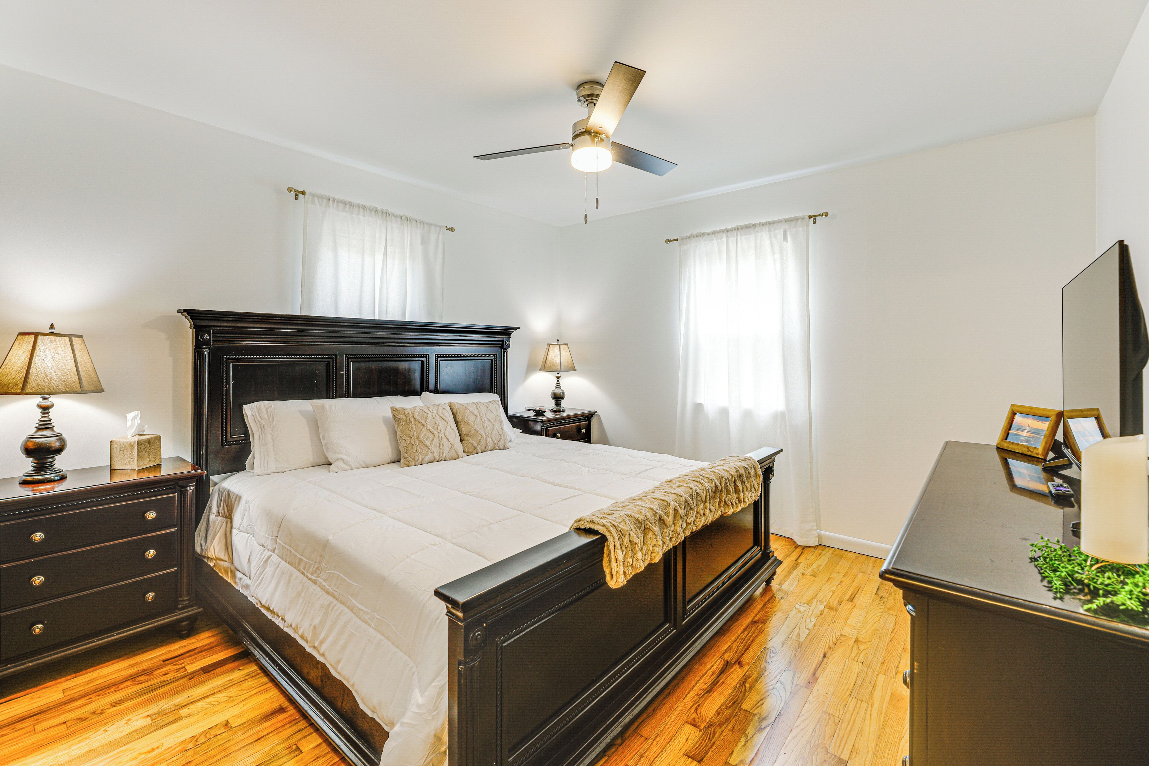 A bedroom with a large bed, wooden floor, ceiling fan, bedside lamps, and a dresser.