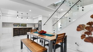 A modern kitchen with white cabinetry, a wooden dining table, and a staircase with glass railings. - Spacious Lidcombe Lux mins to Sydney Olympic Park (Lidcombe)