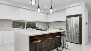 A modern kitchen with white cabinetry, a stainless steel refrigerator, a large island with stools, pendant lighting, and a window with a view of greenery. - Spacious Lidcombe Lux mins to Sydney Olympic Park (Lidcombe)