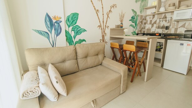 A living room with a beige sofa, two wooden chairs, a small table, and a kitchen area with a microwave, shelves, and a refrigerator. - Beach apartment on the Brazilian northeast coast - Maracaípe. (Ipojuca)