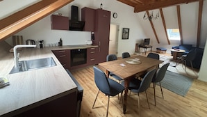 Ein gemütliches Dachzimmer mit einem Holzesstisch, Stühlen, einer Küchenzeile mit Schränken und einem Schlafplatz im oberen Bereich.