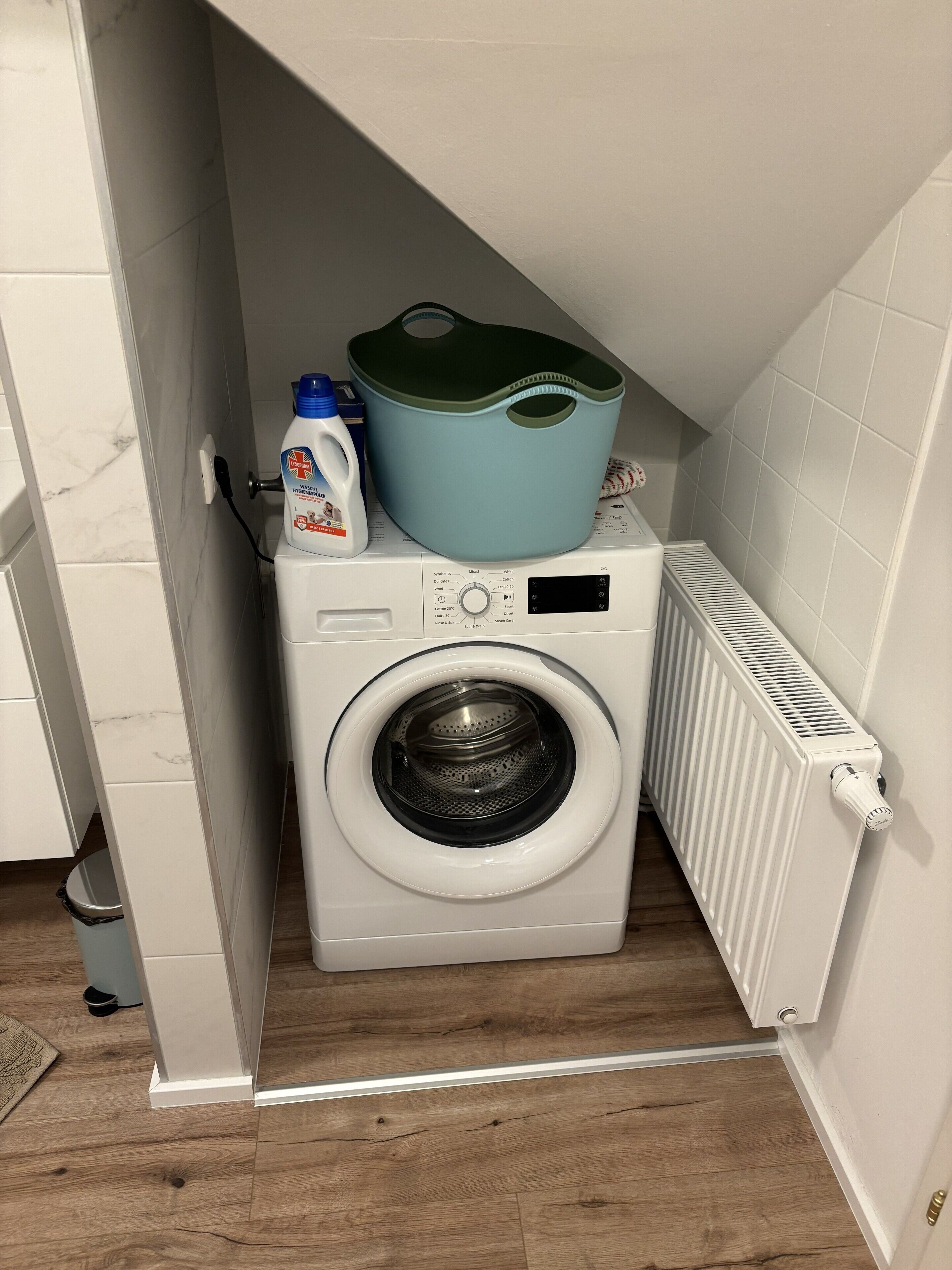 A compact laundry area with a front-loading washing machine, a drying rack, and a radiator.