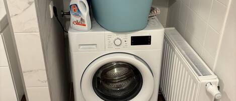 A compact laundry area with a front-loading washing machine, a drying rack, and a radiator.