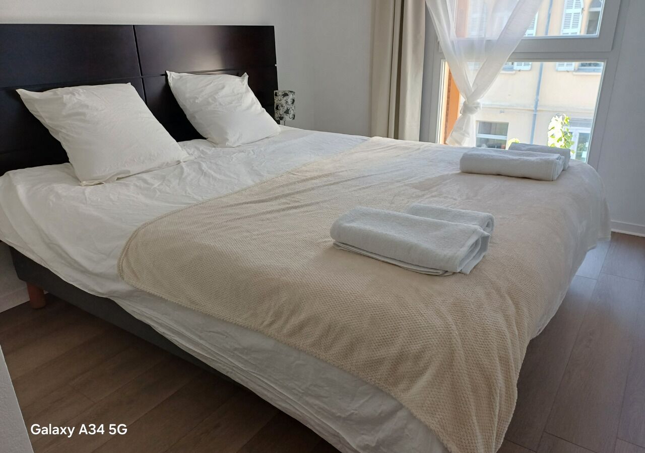A neatly made bed with white linens and a beige blanket, a dark headboard, and a window with curtains.