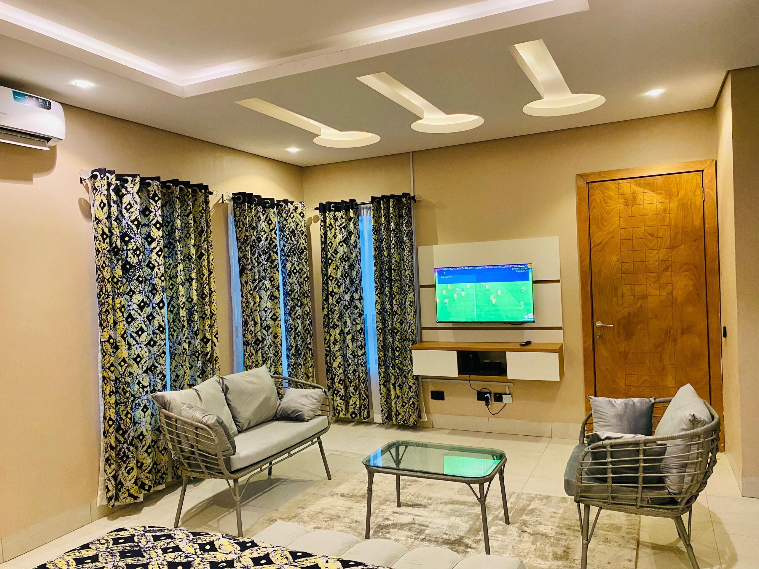 Un salon comprenant un canapé, deux fauteuils, une table basse en verre, des rideaux à motifs, un téléviseur posé sur un meuble en bois et un ventilateur de plafond.