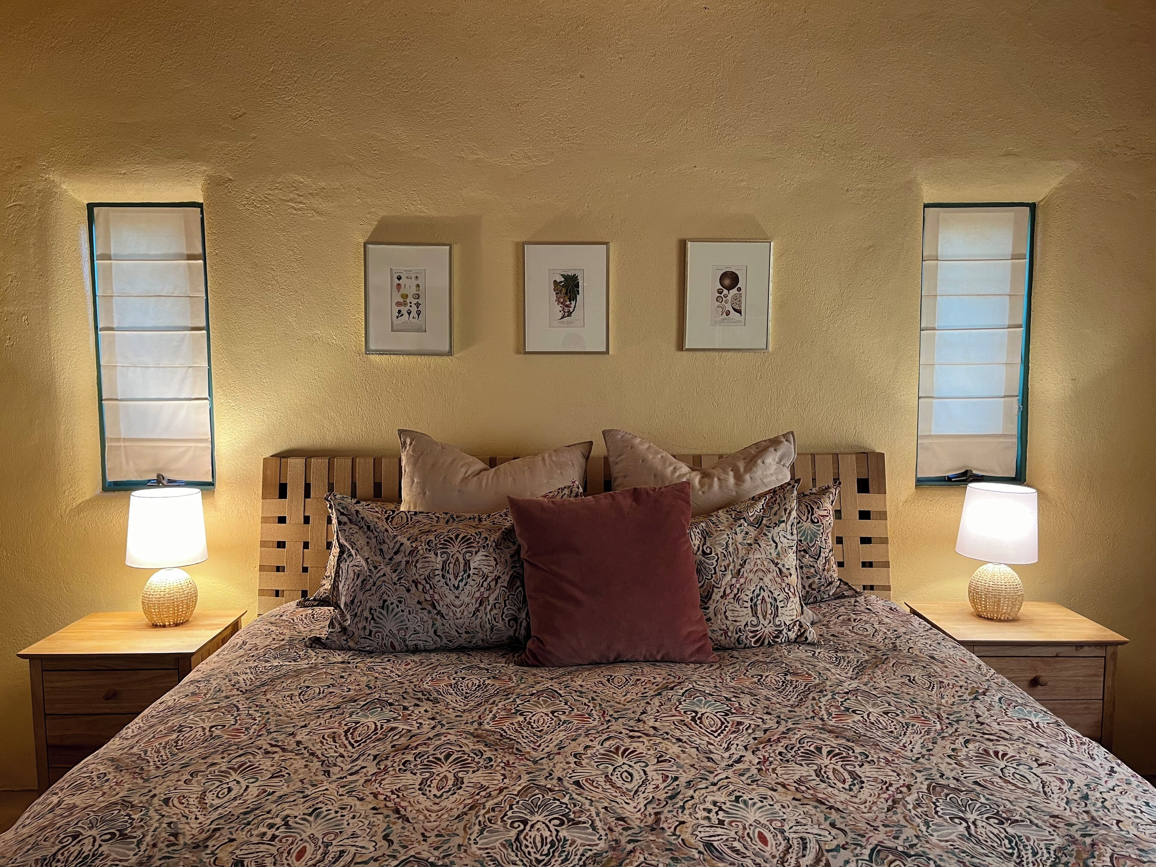 Camera da letto con copriletto decorato, numerosi cuscini, due lampade da comodino e tre quadri appesi al muro.