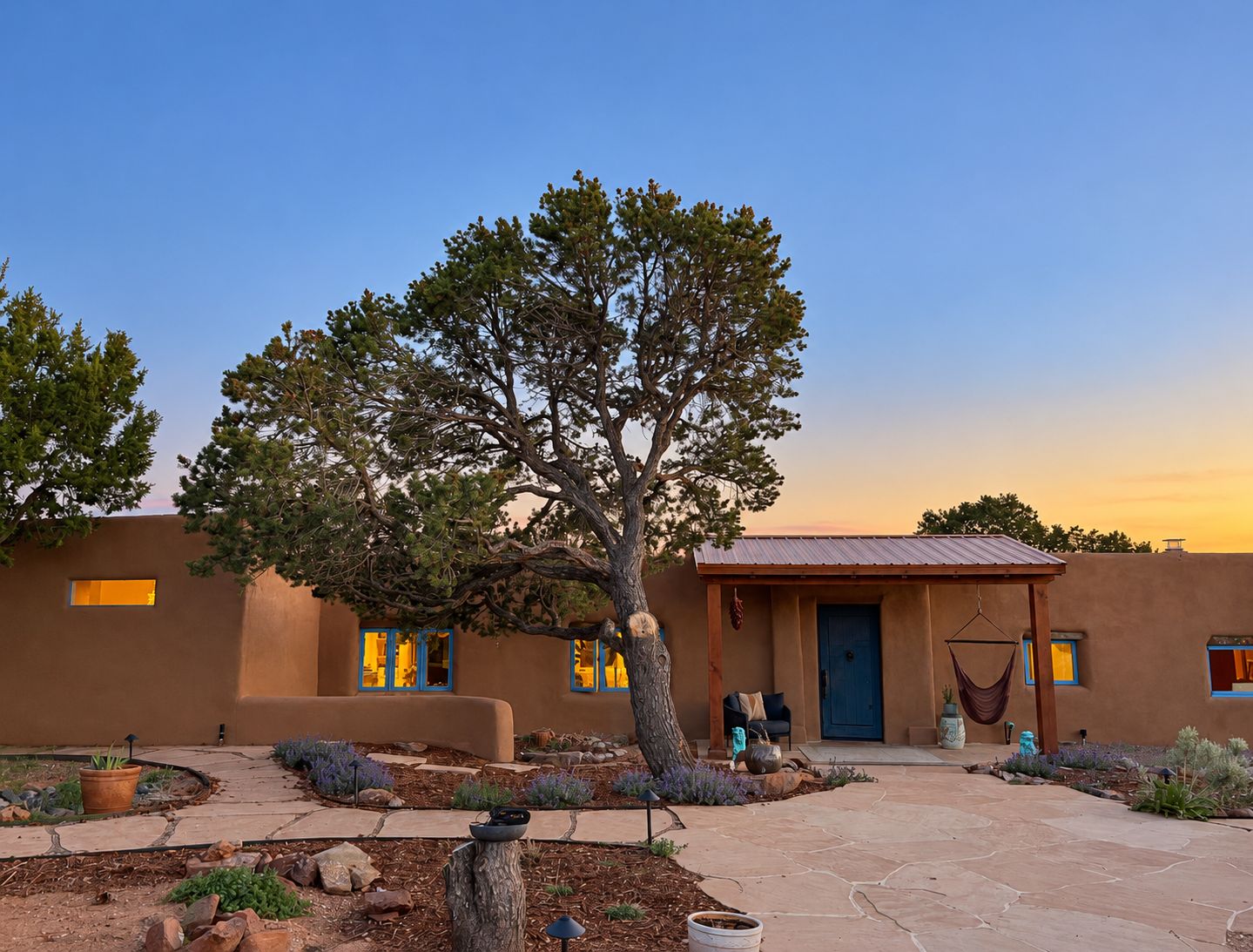 A building with a large tree in the courtyard, a door with a hanging hammock, and a window with blue trim.