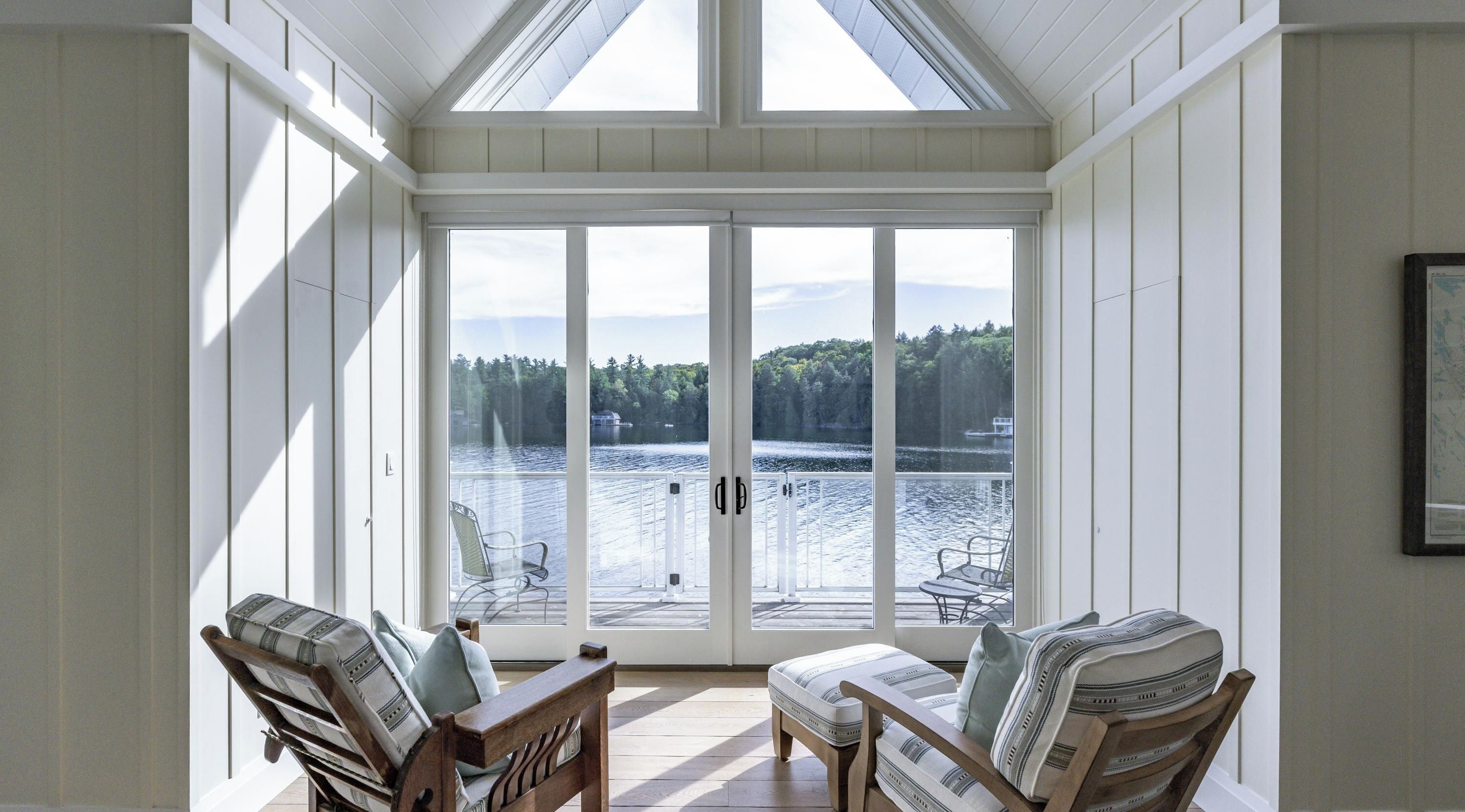 A sunroom with large windows overlooking a lake, furnished with two wooden chairs and a small table.