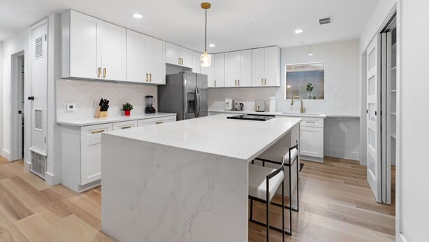 Une cuisine moderne avec des meubles blancs, un grand îlot en marbre, des appareils électroménagers en acier inoxydable et un luminaire suspendu.