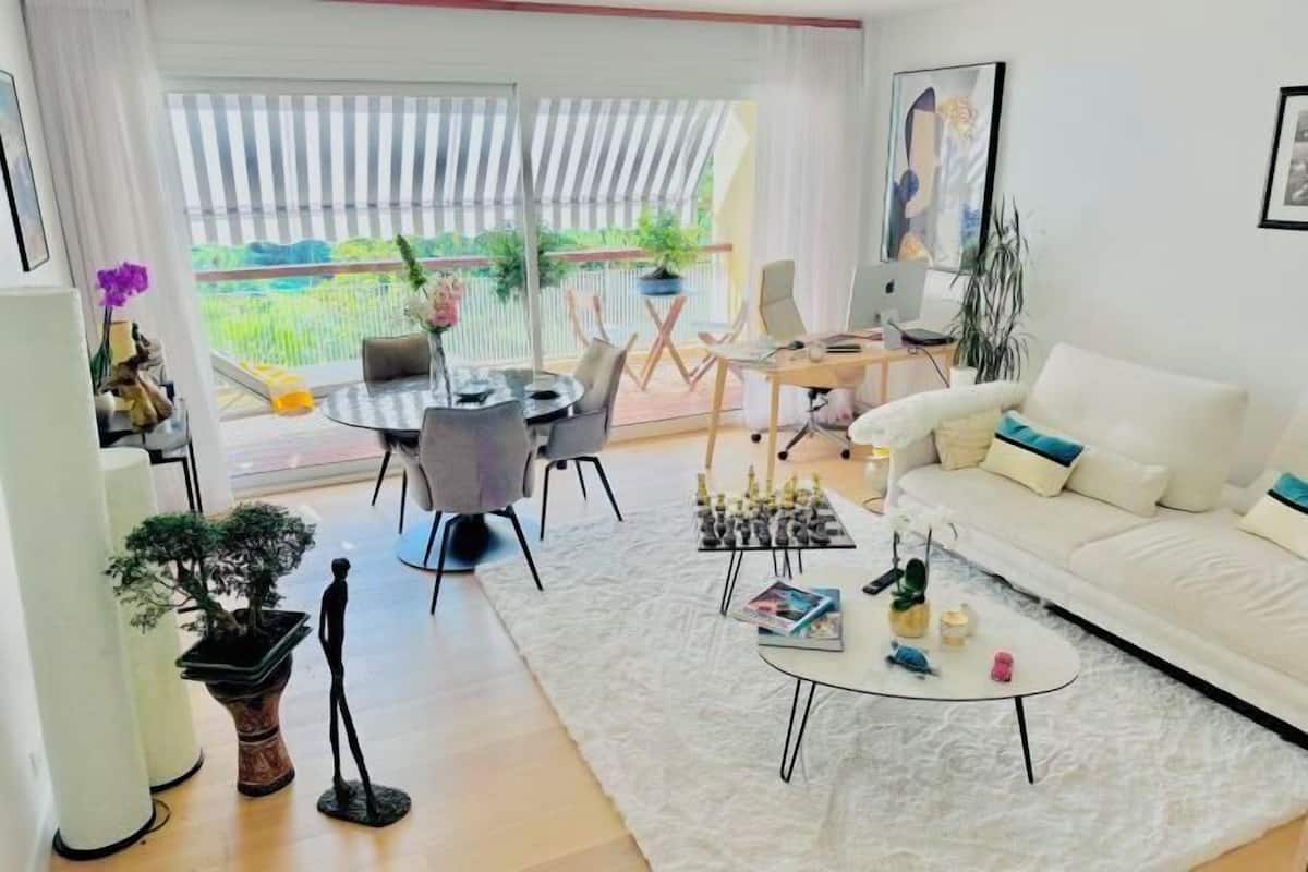 A modern living room with a white sofa, a round coffee table, and a dining area with a table and chairs. There is a balcony with a view of greenery.