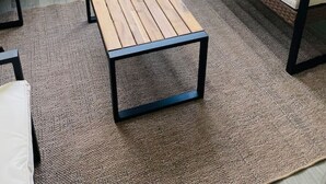 Un balcon avec une table en bois et un banc, un sol recouvert de moquette et une vue sur une rue résidentielle.