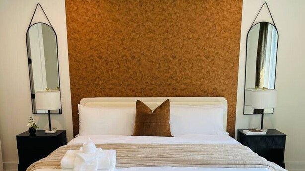 Un grand lit avec du linge de lit blanc, une tête de lit marron, deux tables de chevet avec des lampes et deux fauteuils blancs.