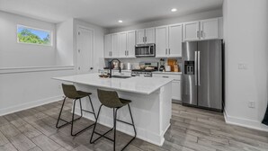 A modern kitchen with white cabinetry, a stainless steel refrigerator, a microwave, and a large island with two bar stools. - Mega Lux Combined Unit - Up to 36 Guests (Austin)