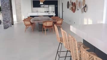 A dining area with a round table and chairs, a kitchen in the background, and a stone wall on the left.