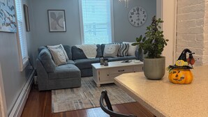 A modern living room with a grey sofa, a coffee table, and a dining area with a bar counter. There is a ceiling fan, a clock on the wall, and a potted plant.