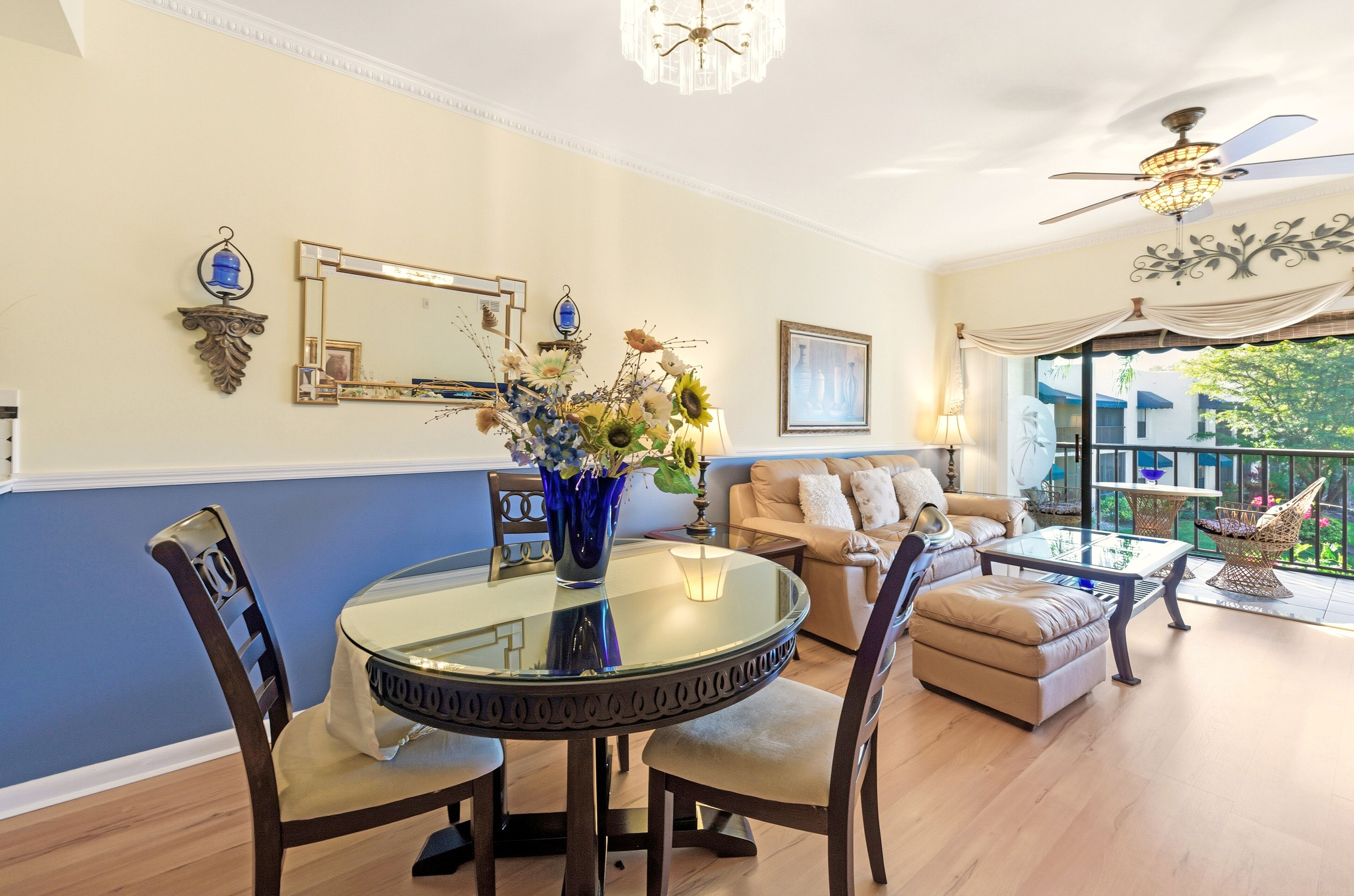 A dining area with a round glass table, chairs, and a vase with flowers. There is a sofa and a balcony with a view.