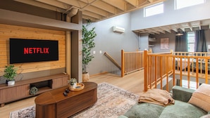 A spacious living area with a large flat-screen TV mounted on a wooden wall, a wooden entertainment unit, a green sofa, a patterned rug, and a staircase leading to an upper level. - 5 mins to Downtown I King I Loft I Keyless Entry (Fargo)