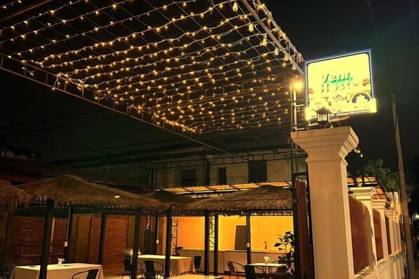 Un espace extérieur aménagé avec des tables et des parasols, une allée couverte illuminée de guirlandes lumineuses et un bâtiment portant une enseigne.