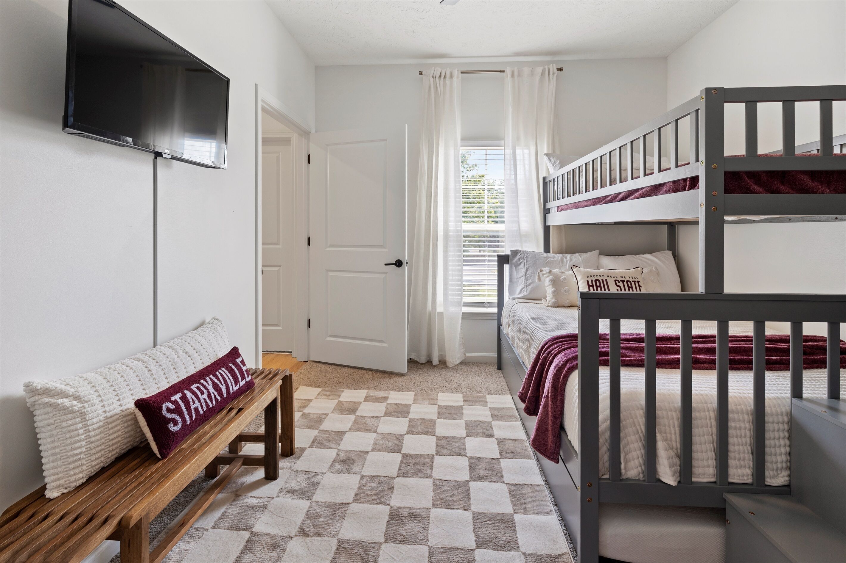 Een slaapkamer met een stapelbed, een enkel bed, een bankje, een televisie en een deur.