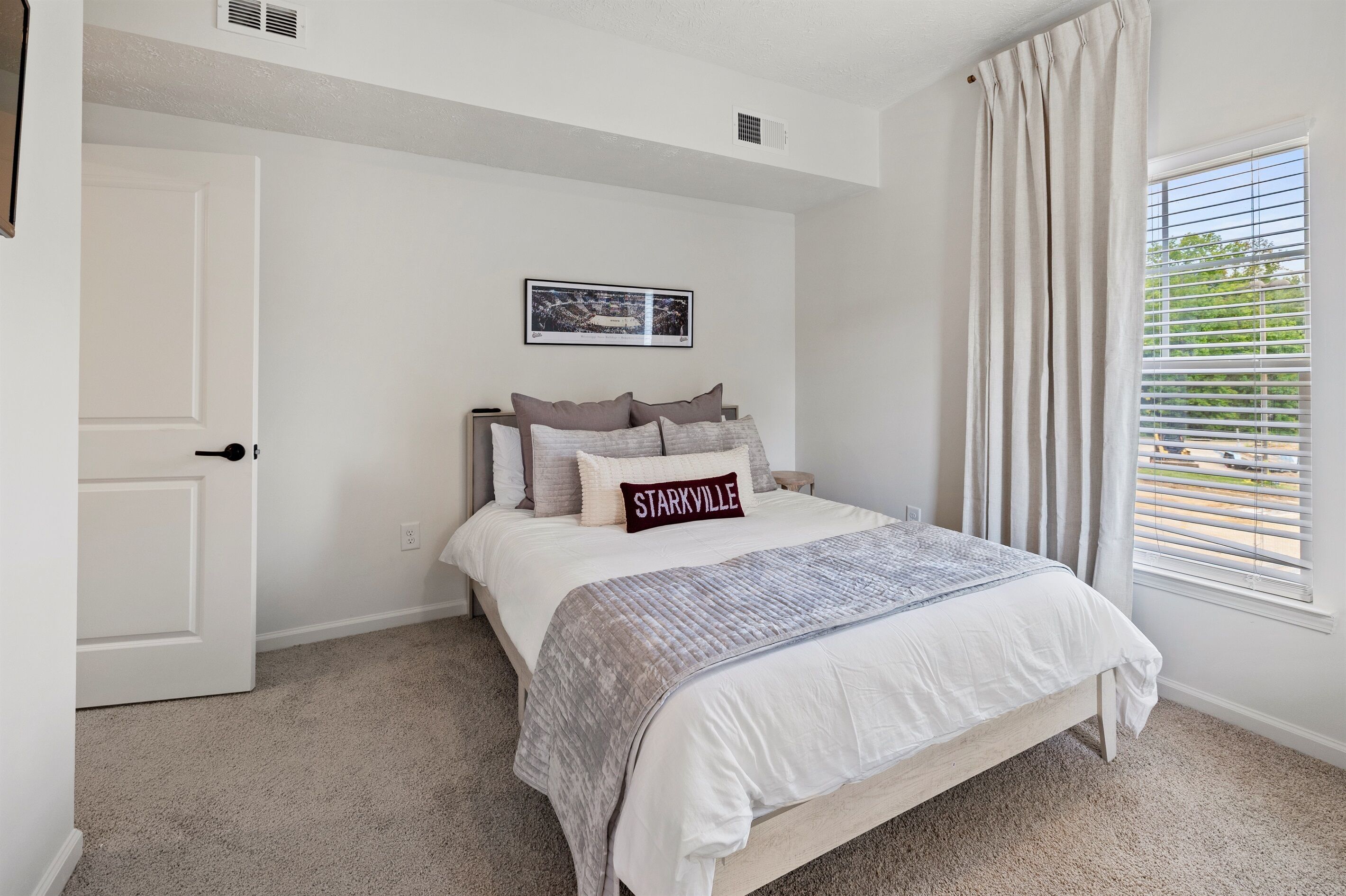 A bedroom with a bed, a door, a window with blinds, and a picture on the wall.