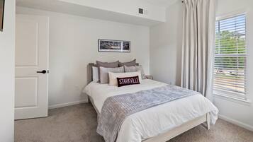 A bedroom with a bed, a door, a window with blinds, and a picture on the wall.