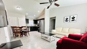 Ein modernes Wohnzimmer mit einem roten Sofa, einem Esstisch mit Stühlen, einer Küche im Hintergrund und einem Deckenventilator.