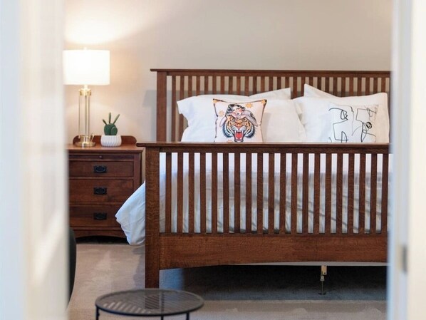 Un dormitorio con una cama de madera, almohadas blancas, una mesita de noche con una lámpara y una pequeña mesa redonda.
