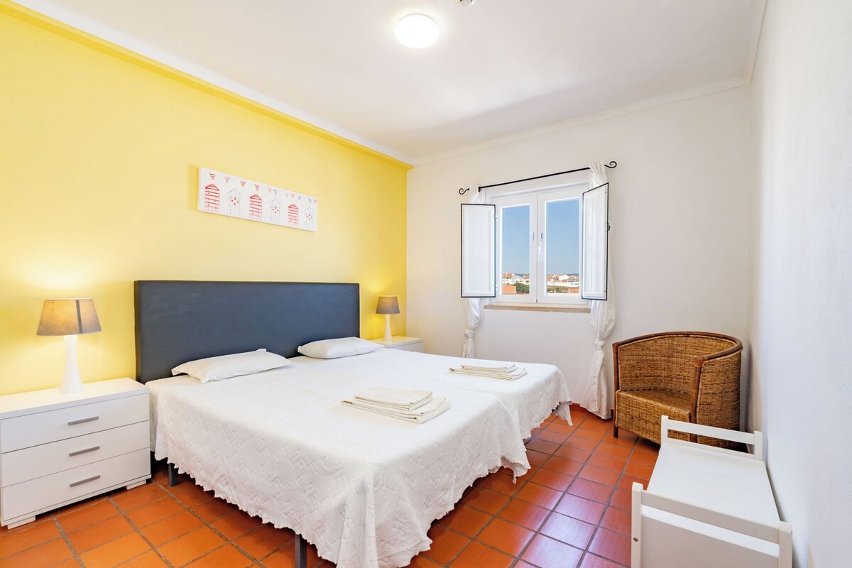 Een hotelkamer met twee bedden, een raam met gordijnen, een stoel en een klein tafeltje.