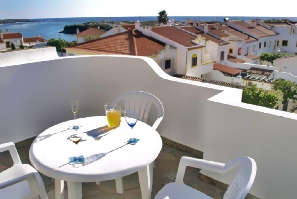 Een ronde tafel met stoelen op een balkon met uitzicht op een kustplaatsje met terracottedaken.