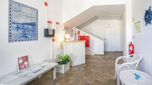 Un espace d’accueil avec un mur carrelé, un comptoir de réception, un banc, des chaises en osier blanc, un extincteur et un escalier.