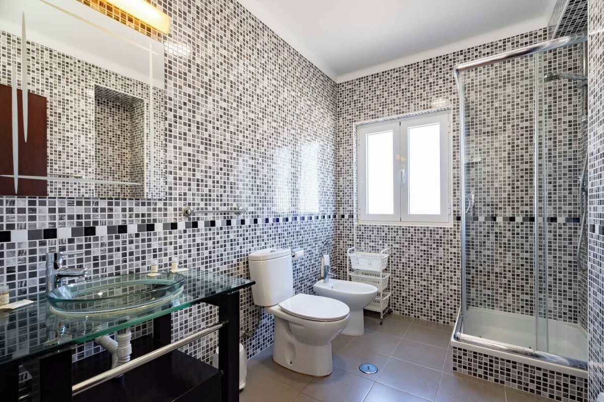 Een badkamer met mozaïektegel wanden, een glazen wastafel, een toilet en een douche.
