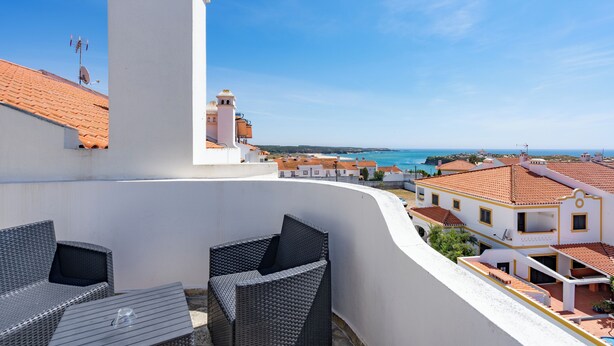 Un balcon agrémenté de meubles en osier, un bâtiment blanc surmonté d’un toit de tuiles rouges, et une vue sur la mer et les autres bâtiments.