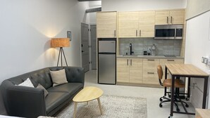 En kompakt opholdsstue med sofa, et lille rundt bord, et tekøkken med mikroovn og køleskab, samt et skrivebord med stol.