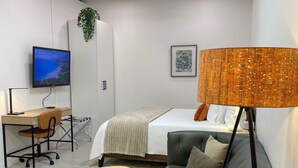 Et moderne, lyst værelse med seng, sofa, skrivebord med computer og fjernsyn.