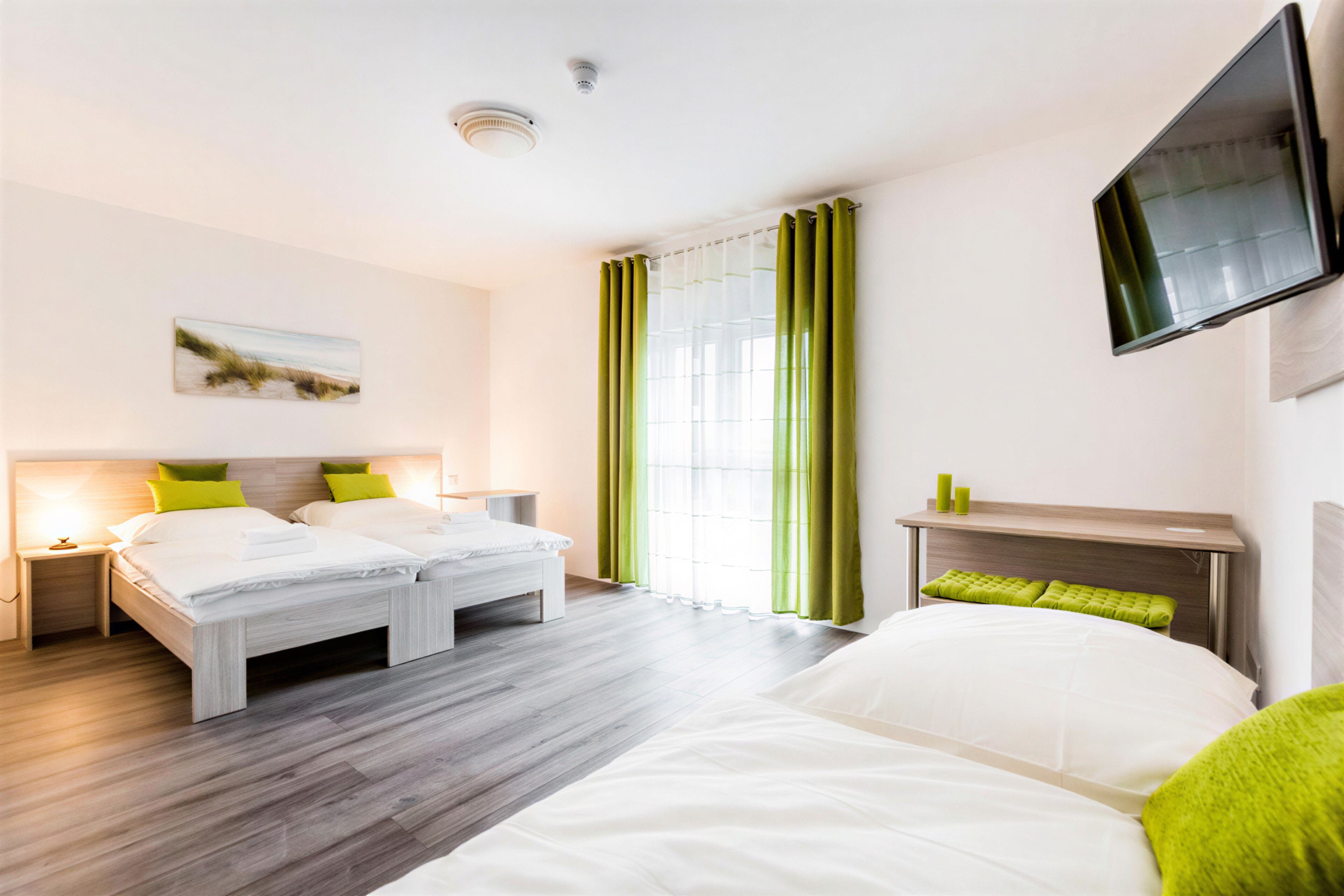 A hotel room with a bed, two bedside tables, a flat-screen TV, a window with green curtains, and a painting on the wall.